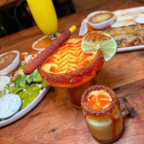 Two vibrant Mexican drinks with chili-lime rims and a lime wedge, alongside a plate of food.