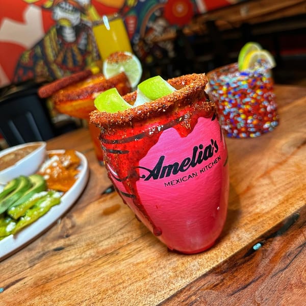 Amelia's Mexican Kitchen's bright pink cocktail with lime and chili rim, alongside appetizers on a wooden table.