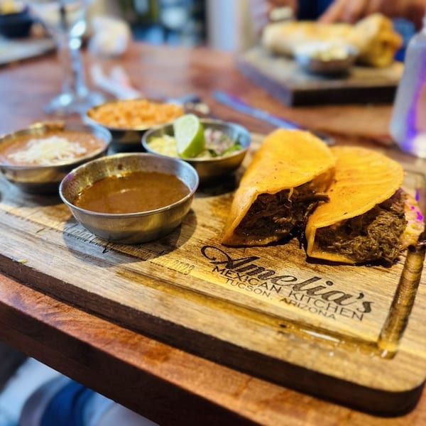Two birria tacos with sides of rice, beans, consommé, and salsa on a wooden board at Amelia's Mexican Kitchen.
