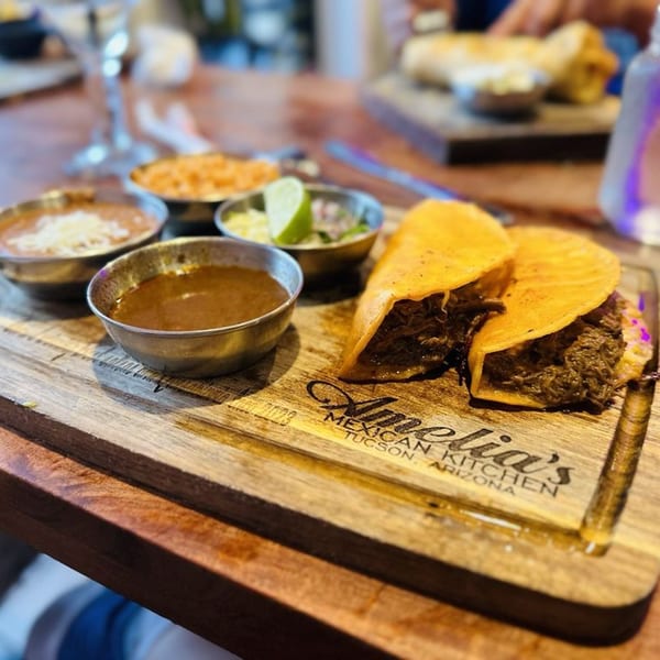 Two birria tacos with sides of rice, beans, salsa, and lime on a wooden board.